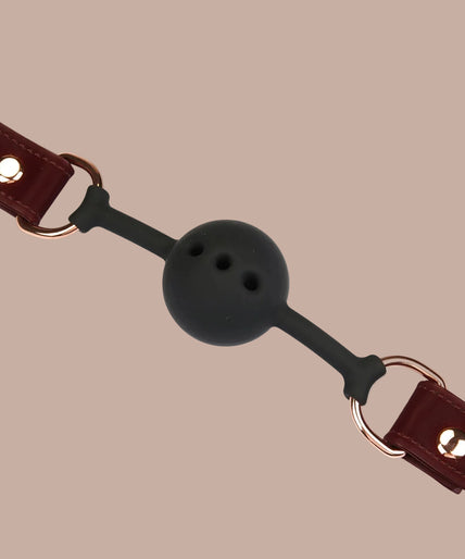 Lieve Ziel Vegan Ball Gag – Metallic Schaduw Donkerrood Dark red ball gag with black perforated ball, metal side rings and adjustable buckle strap, angled view.