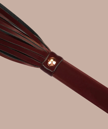 Liebe Seele Vegan Flogger – Metallic Schaduw Donkerrood Dark red flogger with narrow falls, matching wrist loop and rose-gold-tone rivets, curved arrangement.